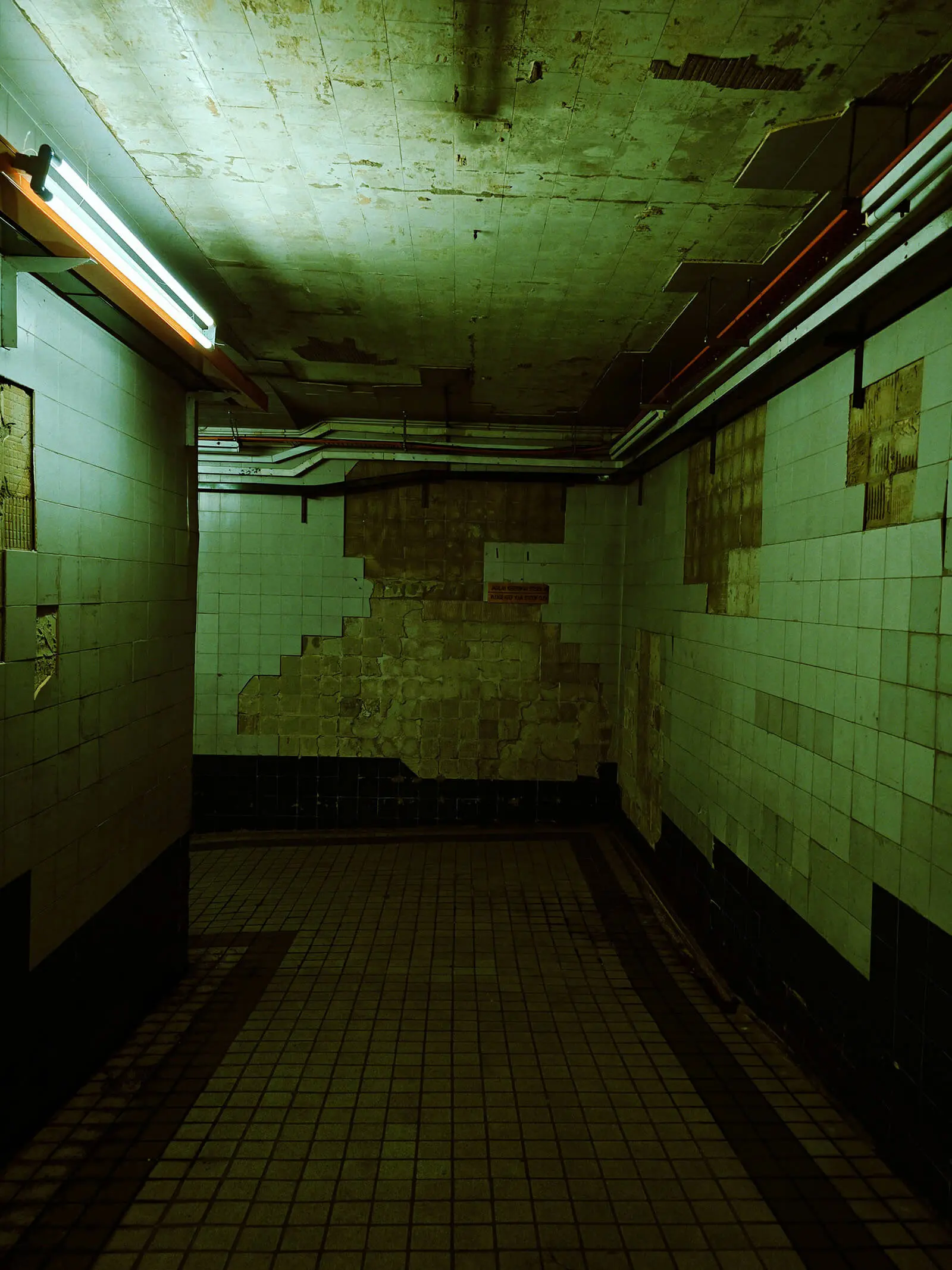 An empty tiled corridor lit by a single green fluorescent tube.